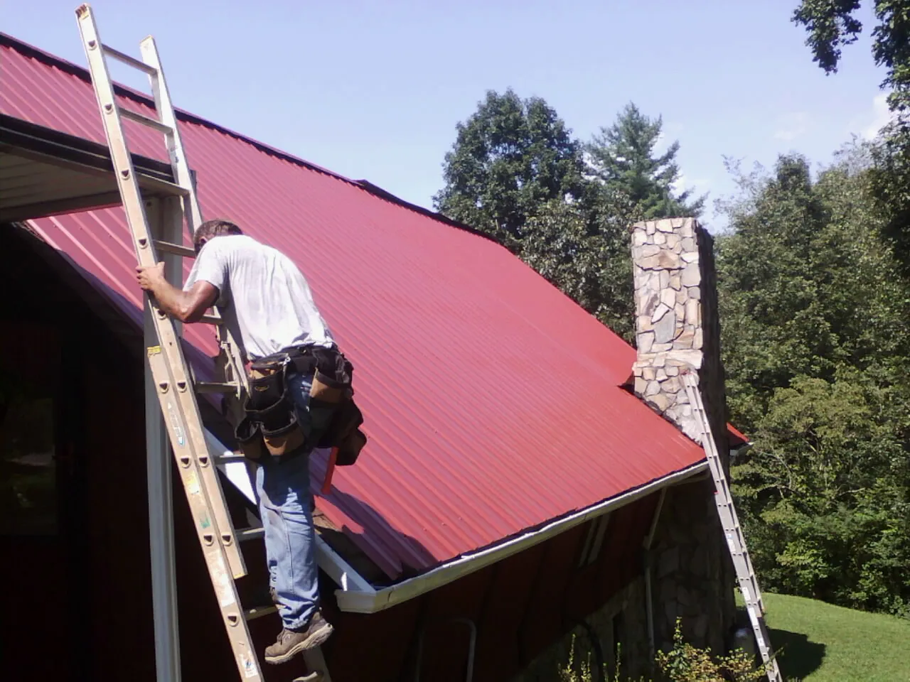 Roofer on a ladder beside a red metal roof and stone chimney — Soffit & Fascia Repair by Roofing Brothers in Kailua